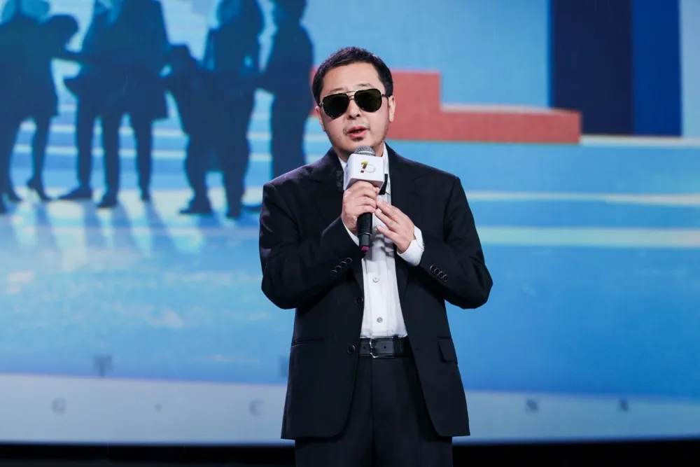 A man in sunglasses and a black suit speaks into a microphone on a blue-lit stage.