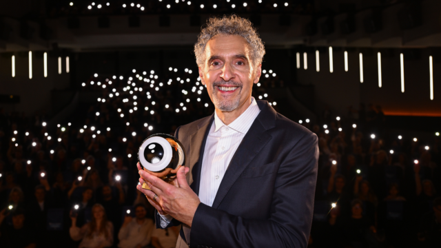 A smiling man in a dark suit holds a trophy before an audience with glowing phone lights.
