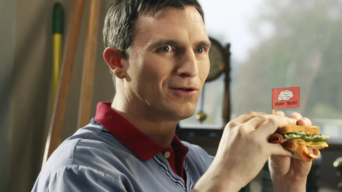 A man in a blue shirt holds a sandwich topped with a small “Brain Trust” flag.