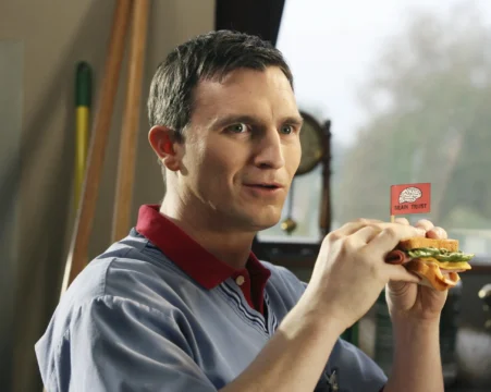 A man in a blue shirt holds a sandwich topped with a small “Brain Trust” flag.