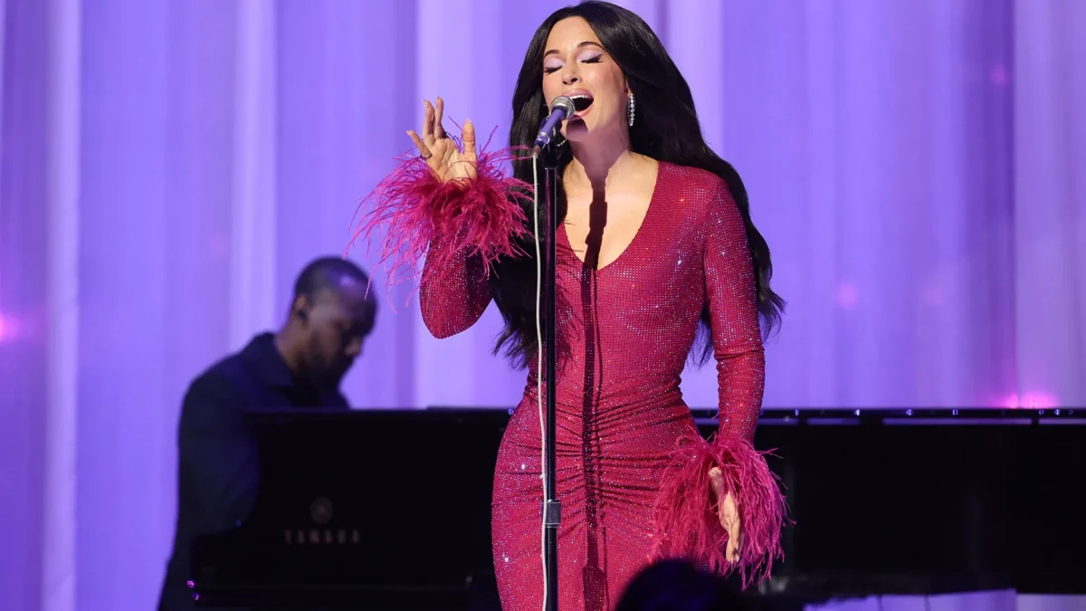 A singer in a sparkling pink feather-trimmed dress performs at a microphone beside a pianist on a purple-lit stage.