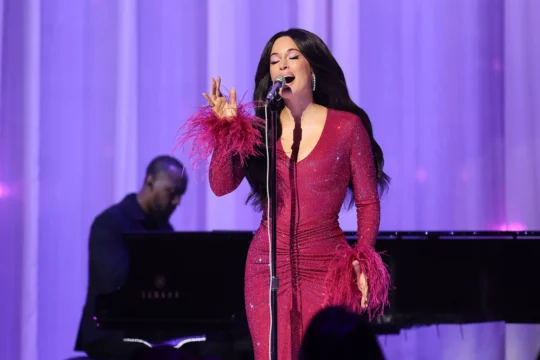 A singer in a sparkling pink feather-trimmed dress performs at a microphone beside a pianist on a purple-lit stage.
