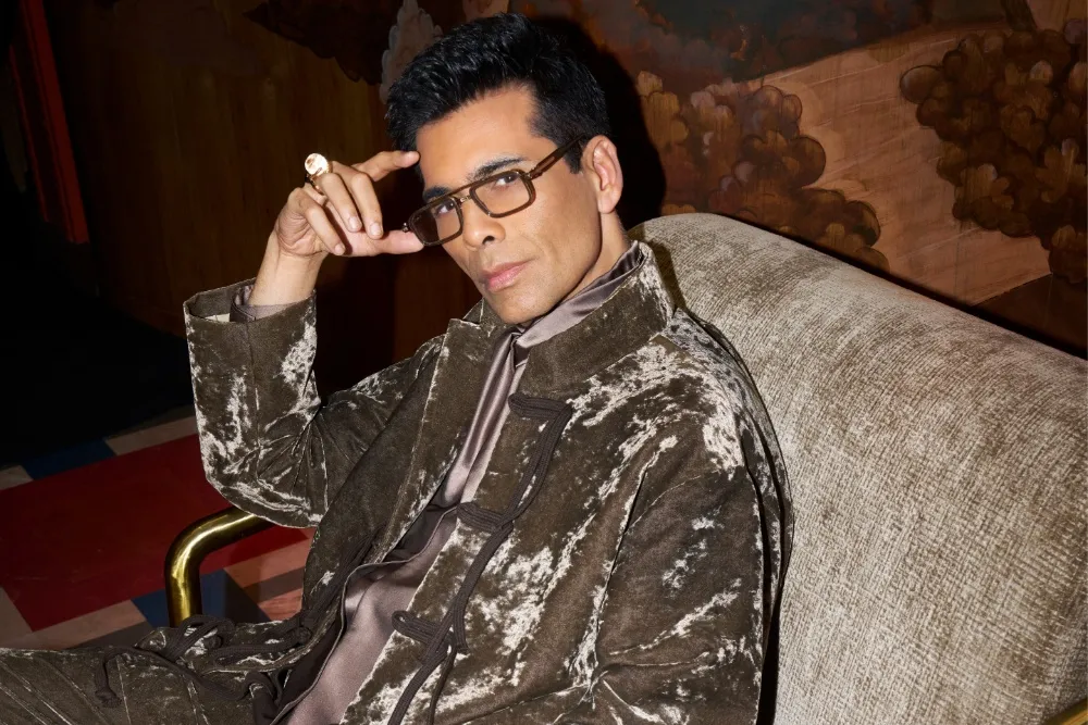 A man in glasses and a velvet jacket sits on a couch with his hand near his temple.