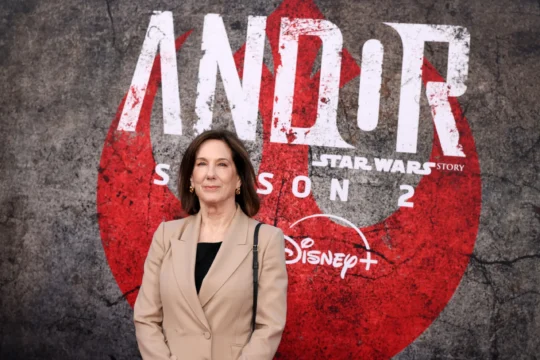 A woman in a tan blazer stands before a large Andor: Star Wars Story Season 2 Disney+ backdrop.