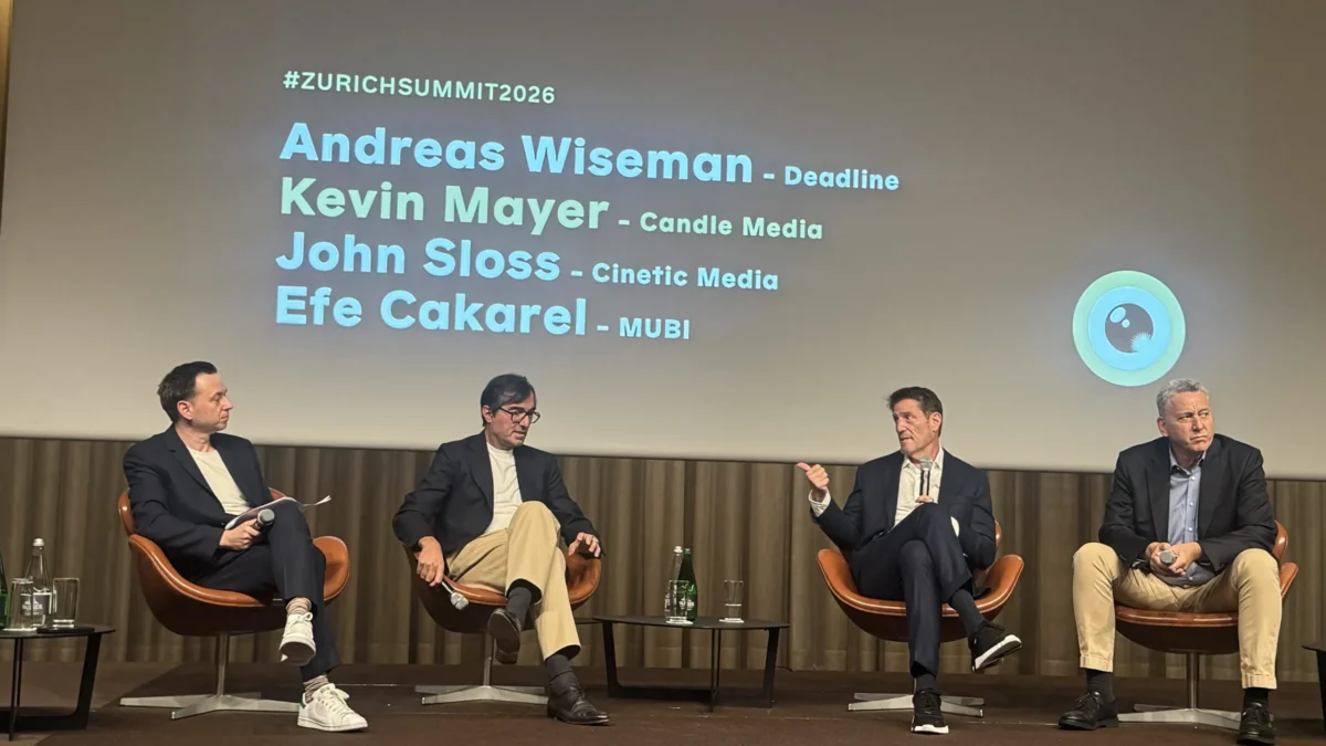 Four people sit onstage with microphones beneath a projected slide listing names and #ZURICHSUMMIT2026.