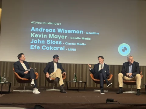 Four people sit onstage with microphones beneath a projected slide listing names and #ZURICHSUMMIT2026.