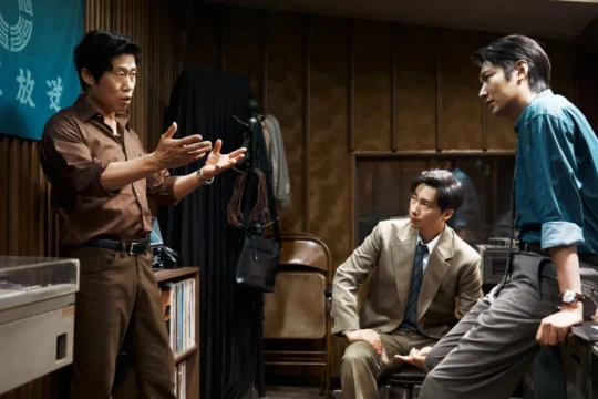 Three men in a wood-paneled room, one gesturing while two listen, with equipment and a Korean banner nearby.