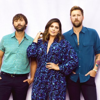 Three adults pose against a light background, the center person in a patterned dress and others in blue shirts.