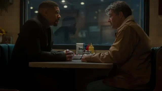 Two men sit across from each other in a dim diner booth, leaning over a table with condiments.