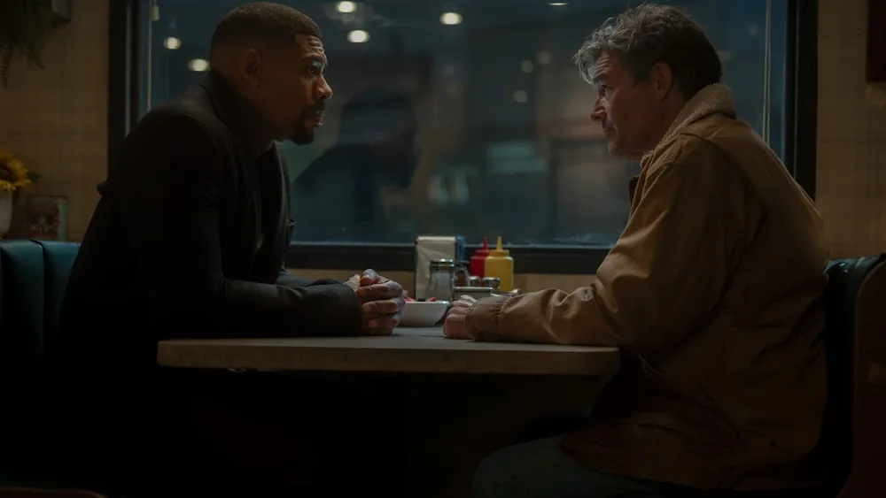 Two men sit across from each other in a dim diner booth, leaning over a table with condiments.
