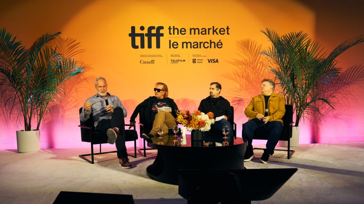 Four men sit on a panel stage beneath a bright orange TIFF The Market backdrop, with plants and microphones.