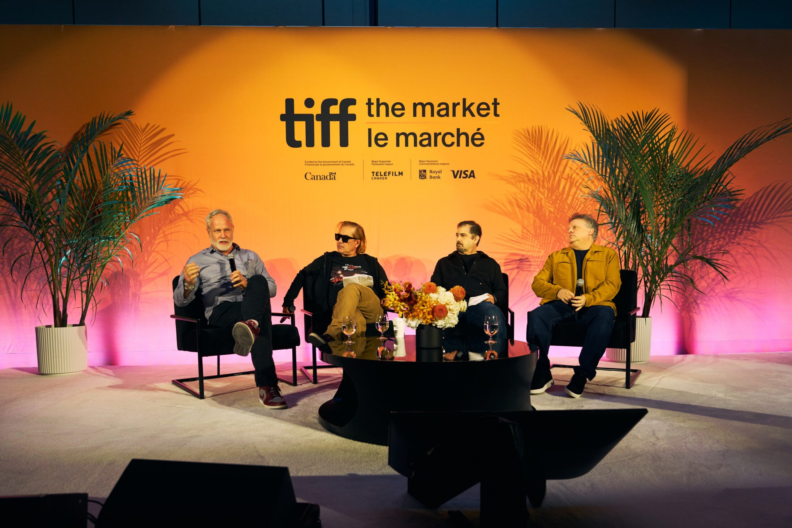 Four men sit on a panel stage beneath a bright orange TIFF The Market backdrop, with plants and microphones.
