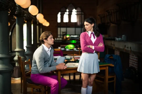 A seated person in bright pink pants looks up at a standing person in a pink cardigan with crossed arms.
