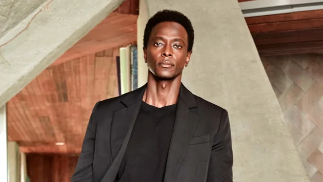 A man in a black blazer and shirt stands before angular concrete and wood architectural elements.