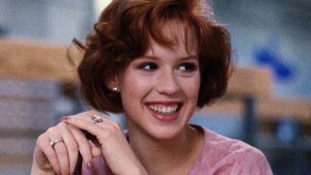 A smiling red-haired young woman in a pink top clasps her hands in a blurred indoor setting.