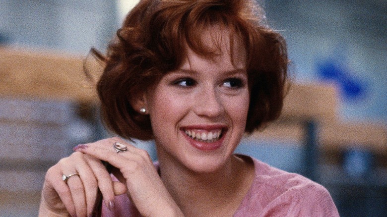 A smiling red-haired young woman in a pink top clasps her hands in a blurred indoor setting.
