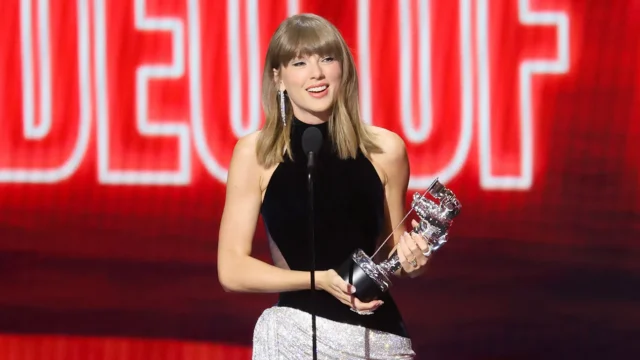 A woman in a black top holds a silver trophy at a microphone before a red backdrop.