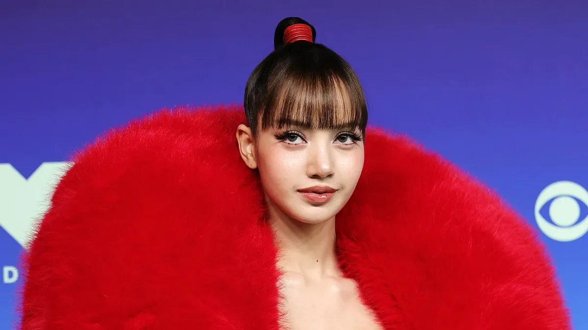 A woman with blunt bangs wears a voluminous bright red faux fur wrap against a blue-purple backdrop.