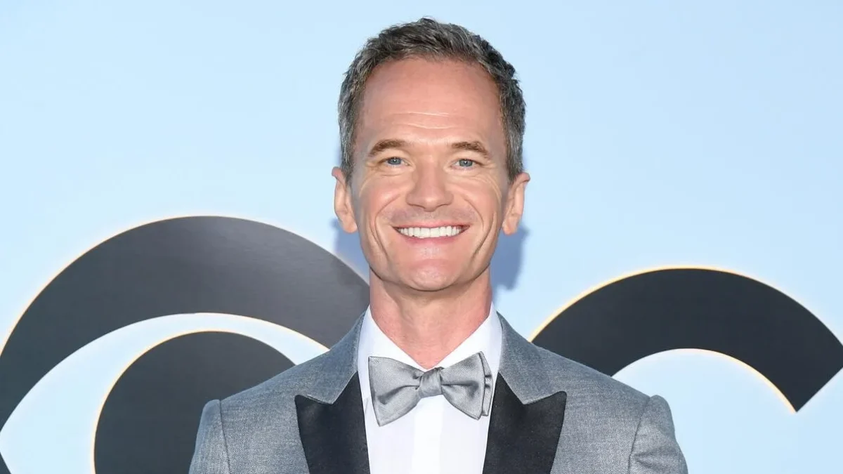Smiling man in a gray tuxedo and bow tie stands before a pale blue backdrop with black circular shapes.