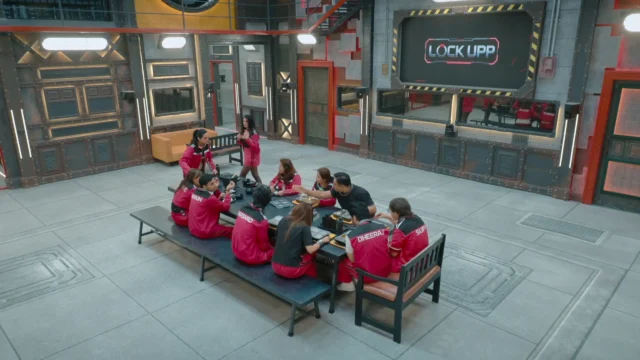 People in red uniforms sit around a table in an industrial room with a large “Lock Upp” screen.