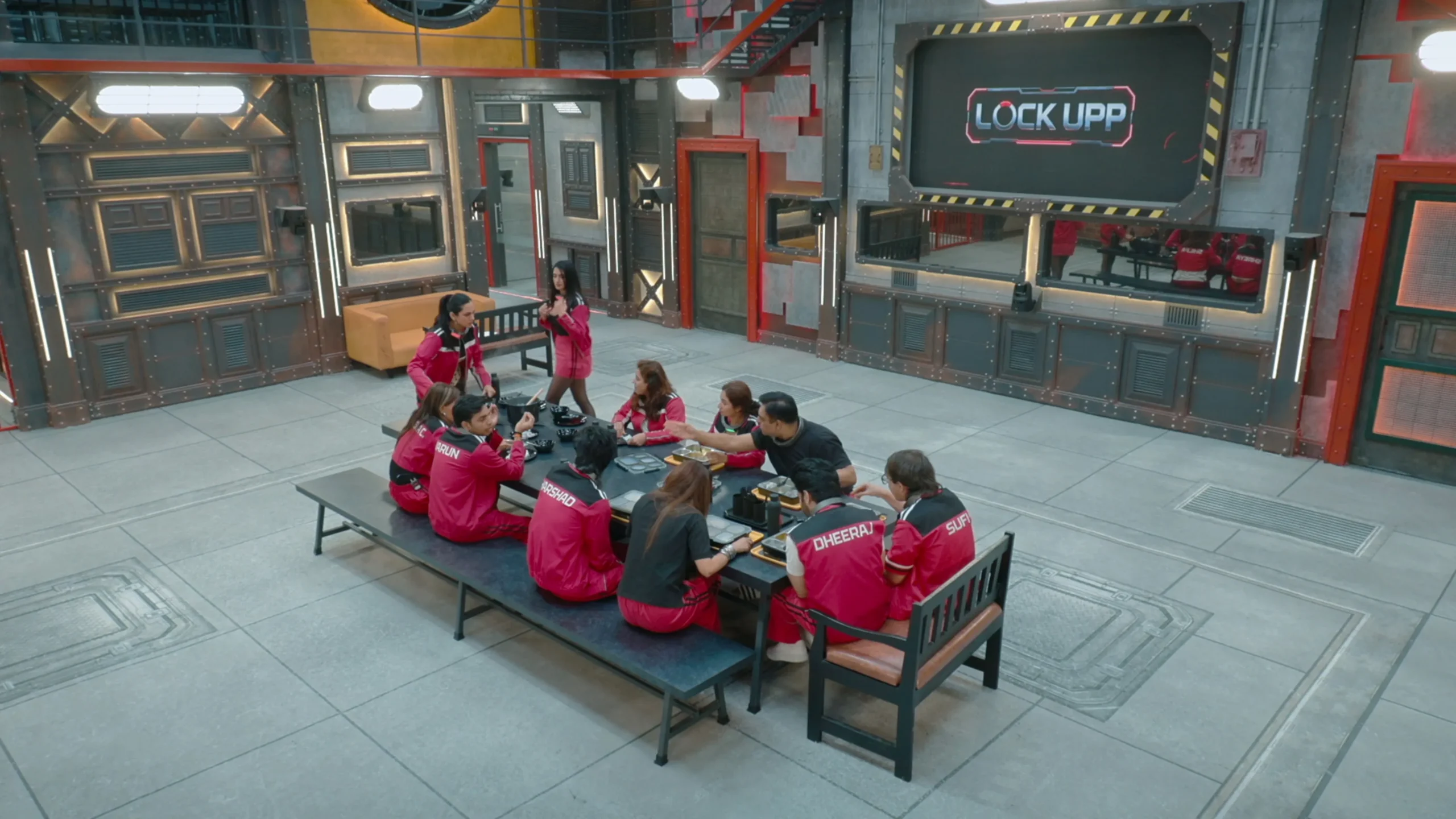 People in red uniforms sit around a table in an industrial room with a large “Lock Upp” screen.