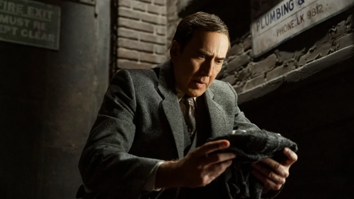 A man in a gray coat holds a dark object beneath a worn plumbing sign.