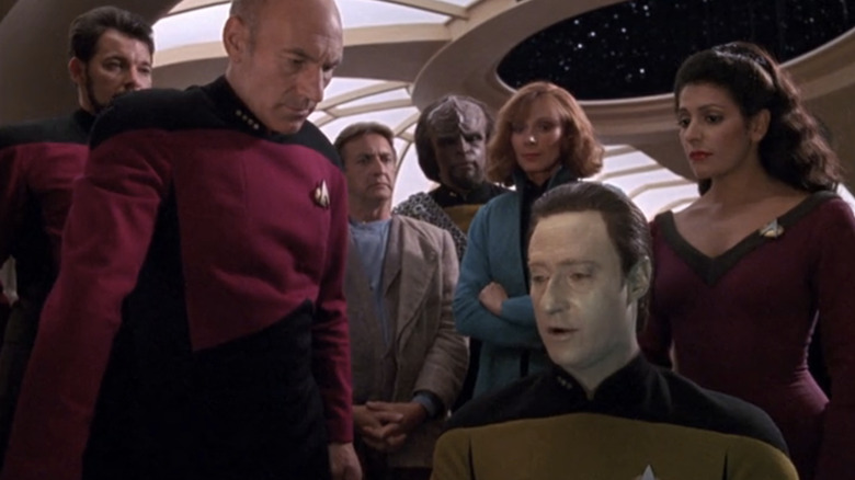 A group in futuristic uniforms gathers around a pale-faced seated man in a starry, windowed room.