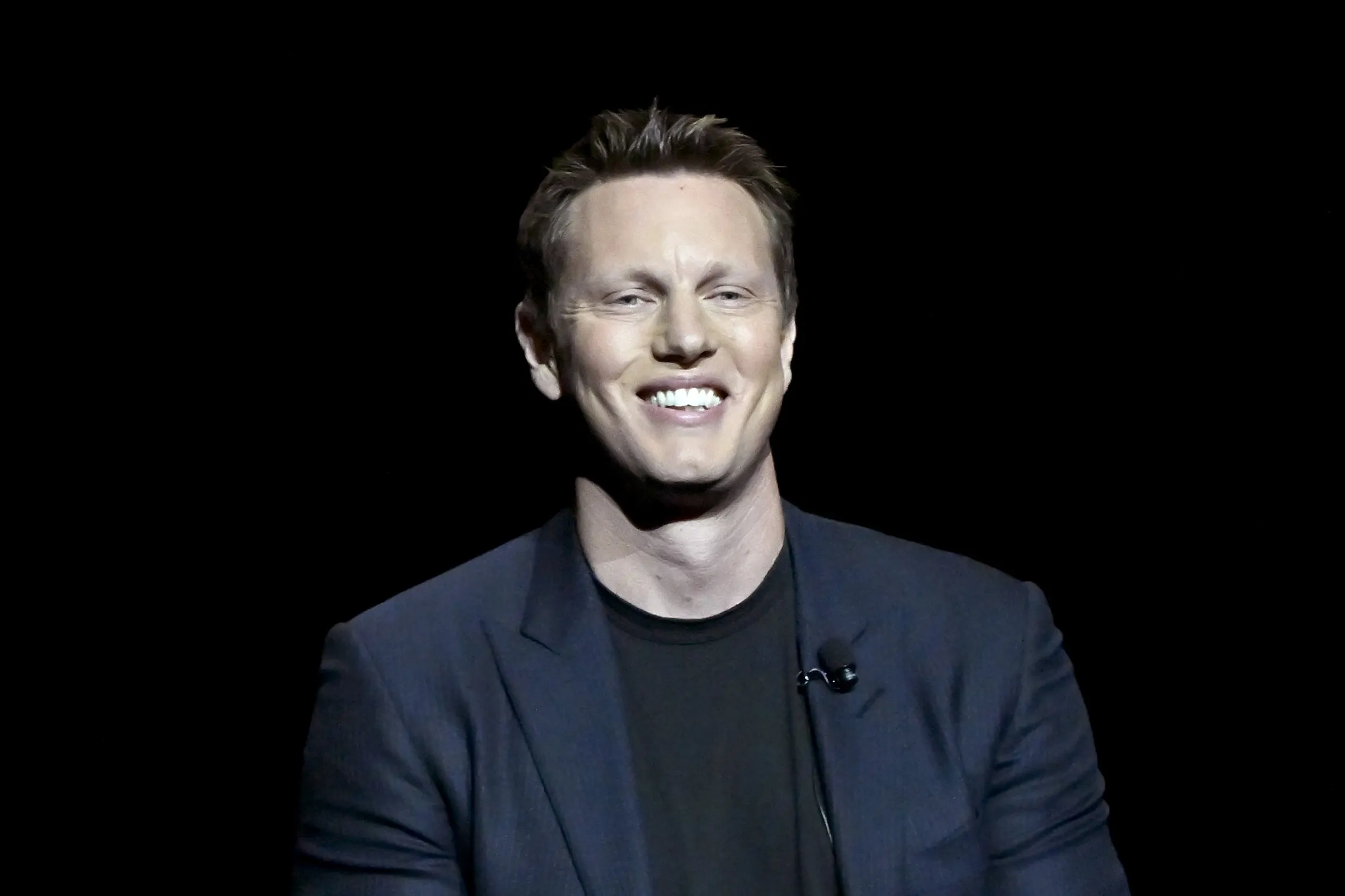 A smiling man in a dark blazer and black shirt sits against a black background.