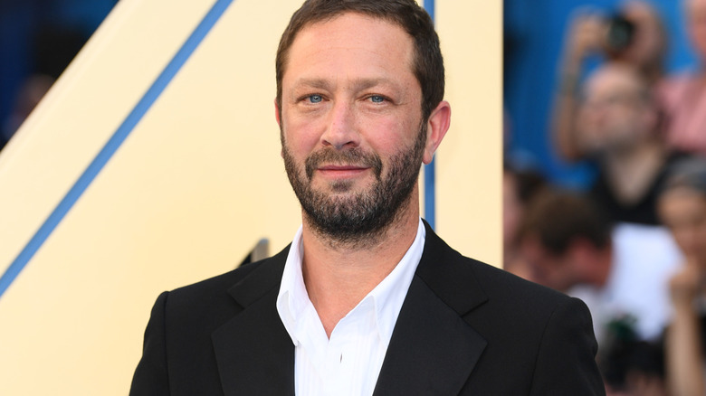 Bearded man in a black suit and white shirt poses against a blurred event backdrop.