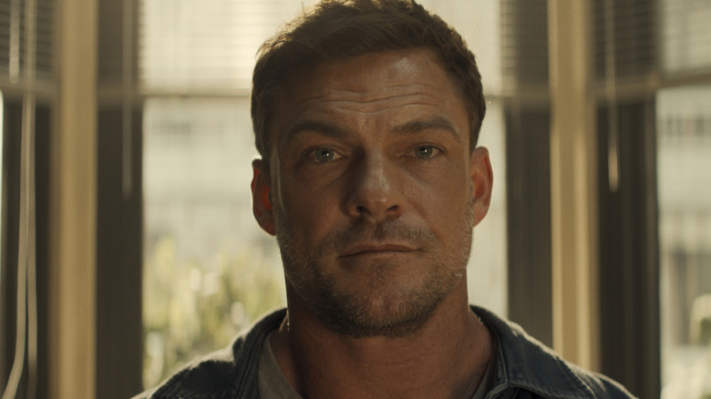 A close-up of a man with short hair and stubble standing in front of sunlit windows.