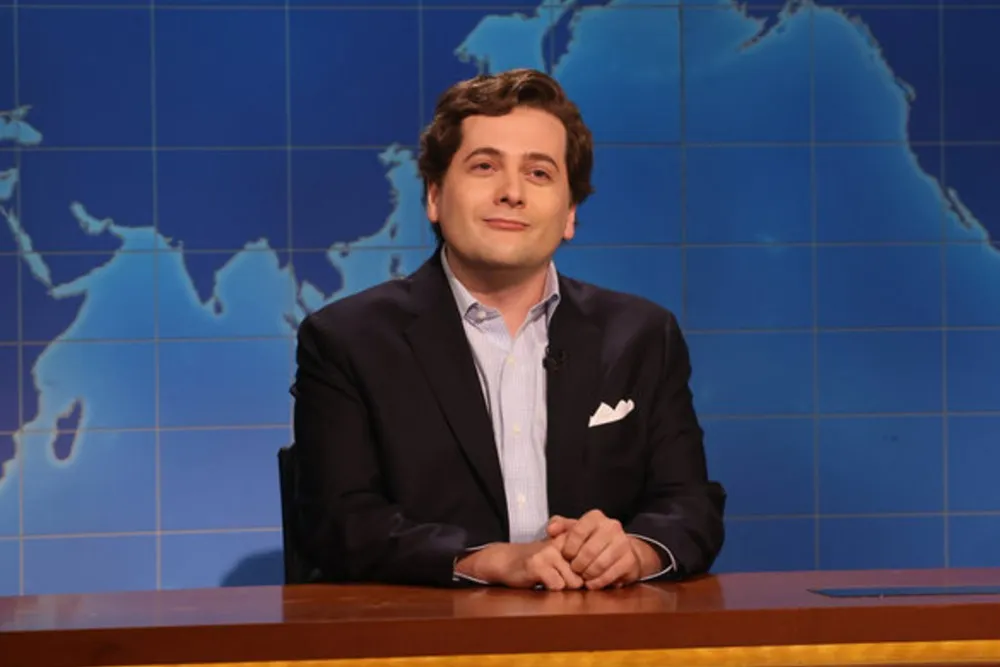 A man in a dark suit sits at a desk before a blue world map backdrop.
