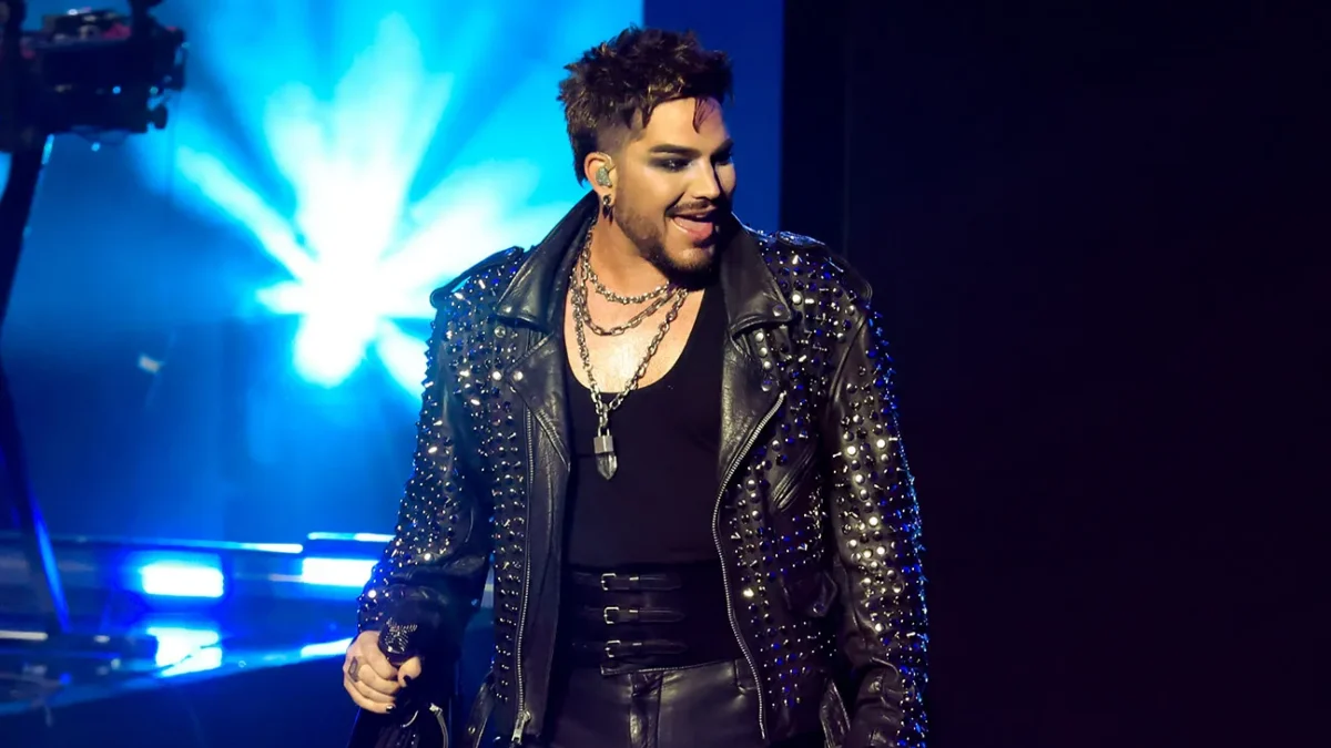 A performer in a studded black leather jacket holds a microphone under bright blue stage lights.