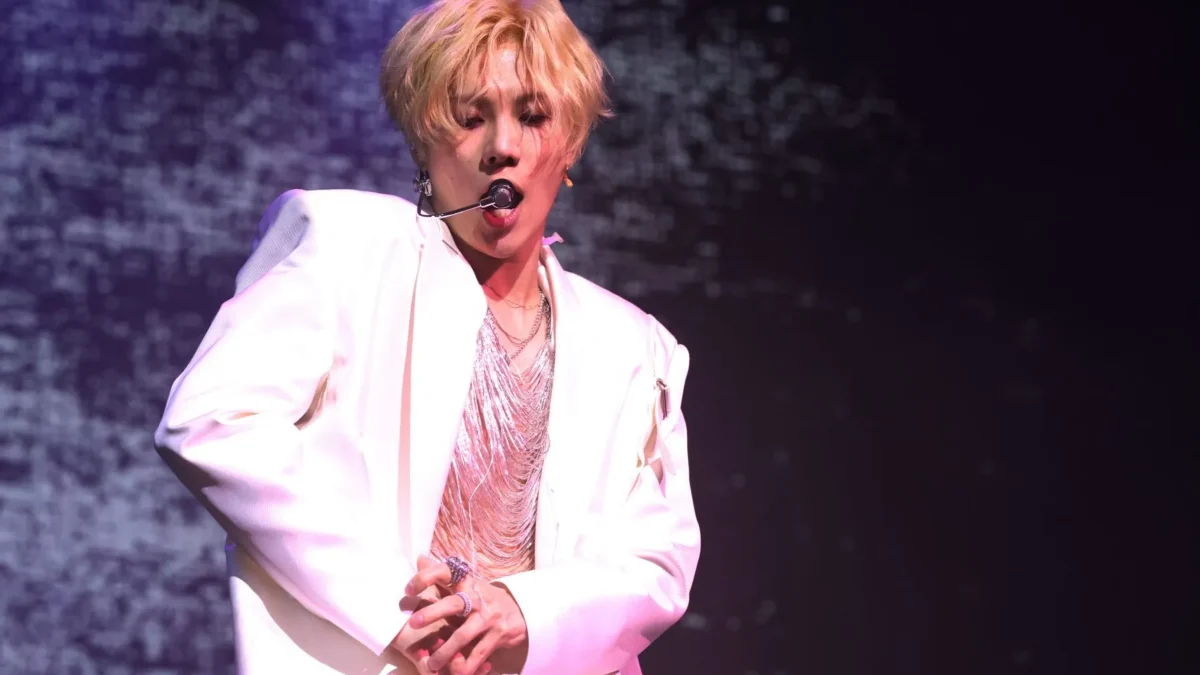 Blond performer in a white outfit sings into a headset microphone against a dark purple stage backdrop.