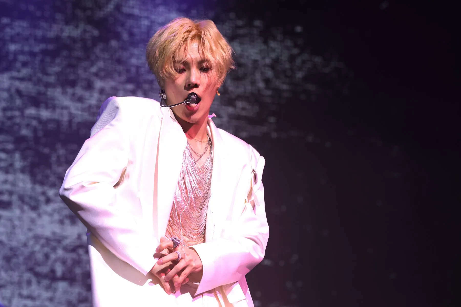 Blond performer in a white outfit sings into a headset microphone against a dark purple stage backdrop.
