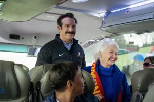 A mustached man in a jacket smiles beside an older woman wearing a blue coat and red scarf.