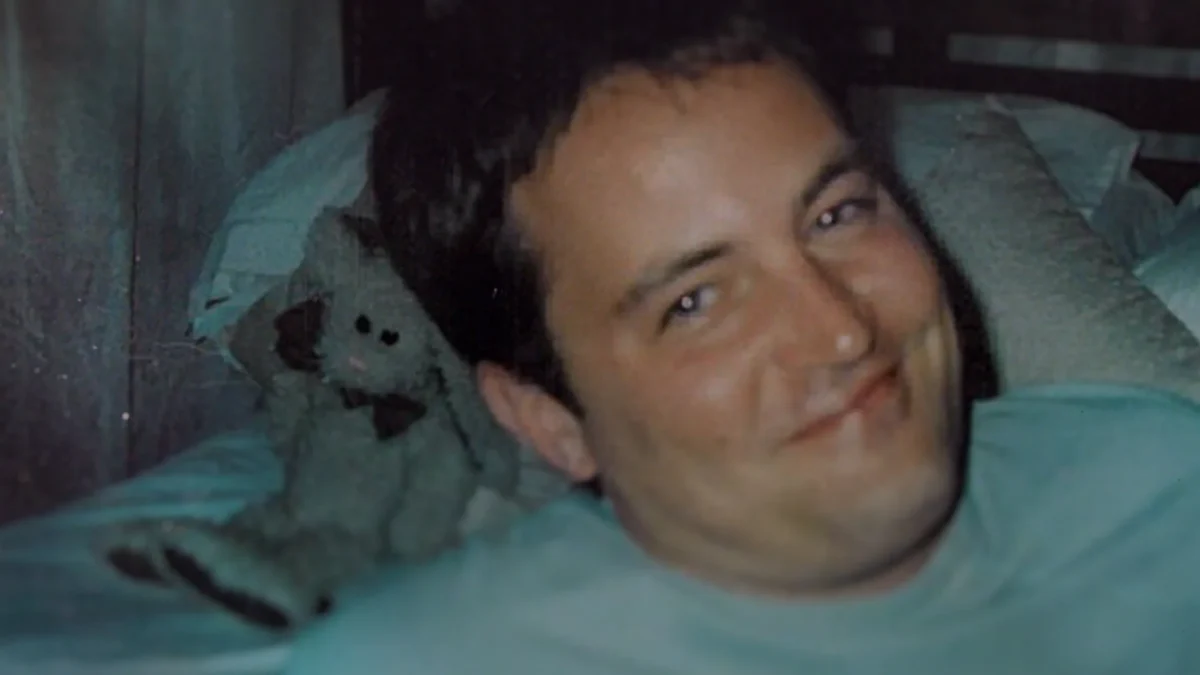A smiling man lies on a bed beside a worn stuffed animal in a grainy, dimly lit close-up.