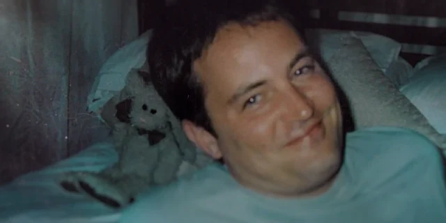 A smiling man lies on a bed beside a worn stuffed animal in a grainy, dimly lit close-up.