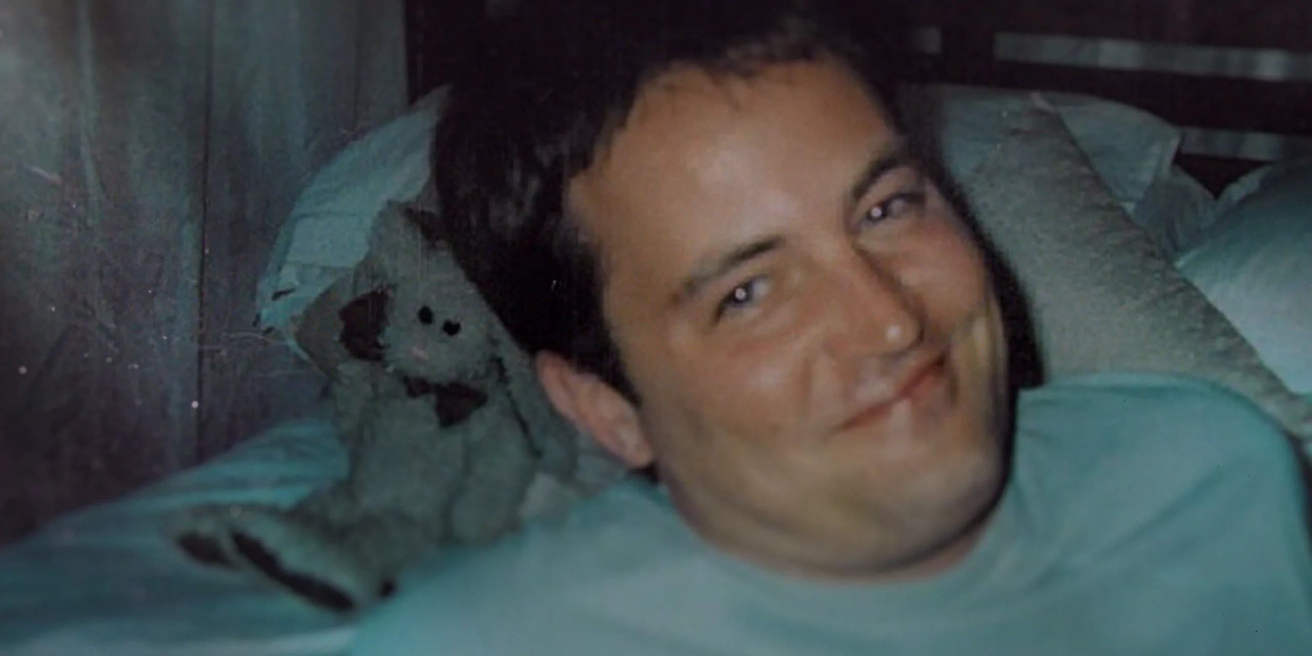 A smiling man lies on a bed beside a worn stuffed animal in a grainy, dimly lit close-up.