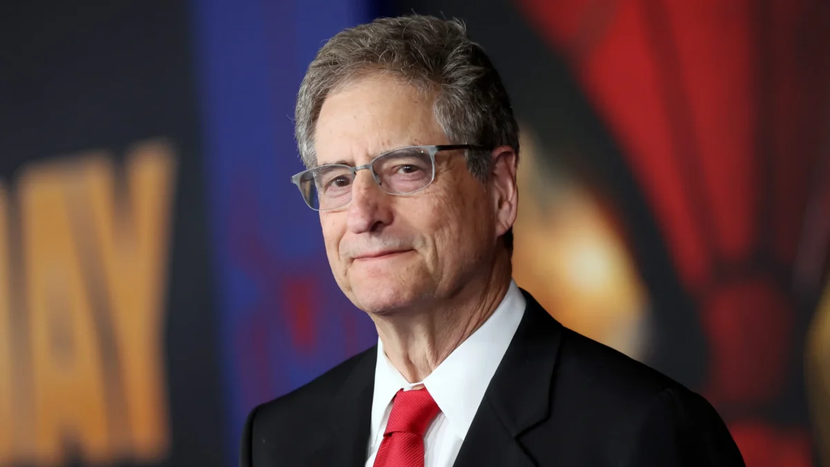 A man in glasses, a black suit, and a red tie stands before a blurred colorful backdrop.