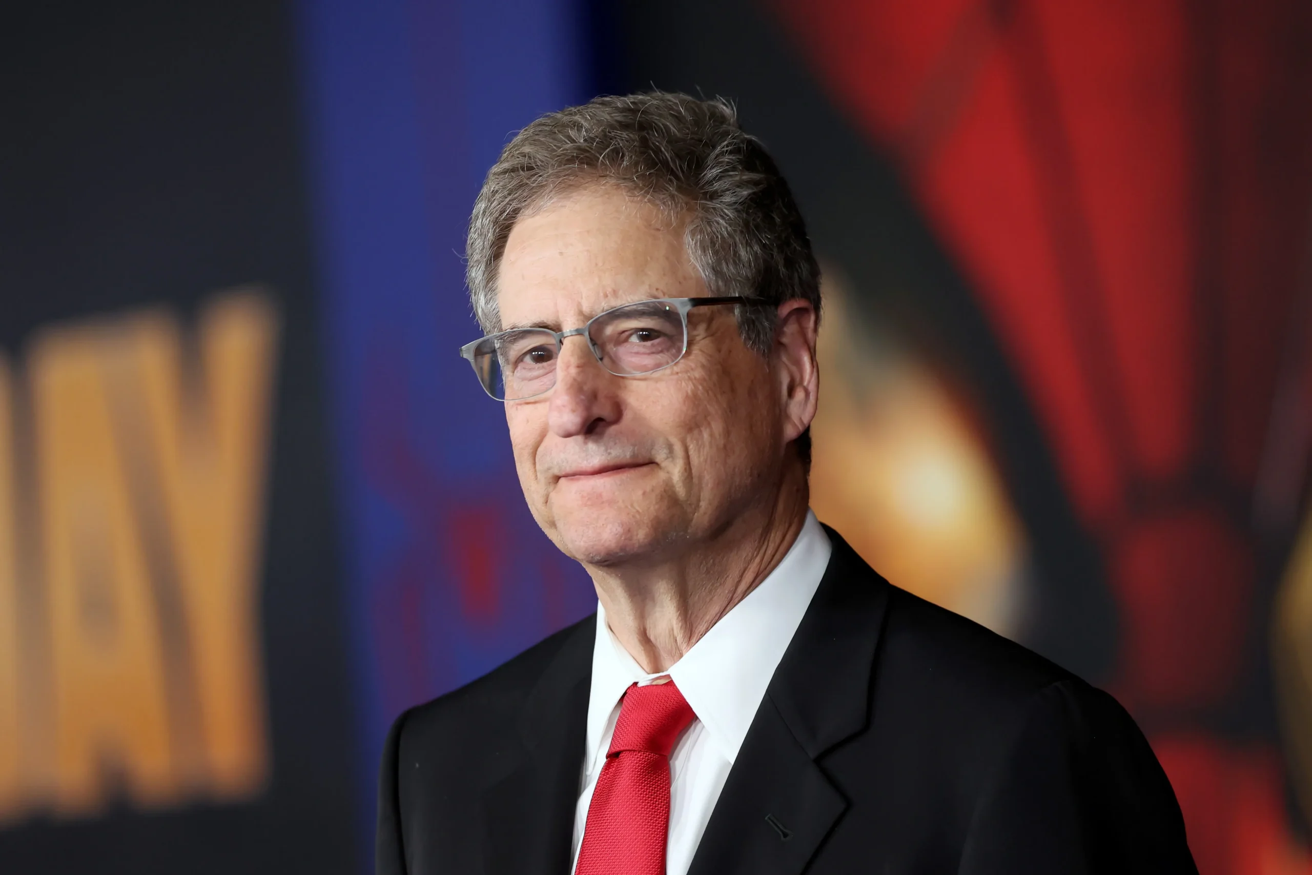 A man in glasses, a black suit, and a red tie stands before a blurred colorful backdrop.