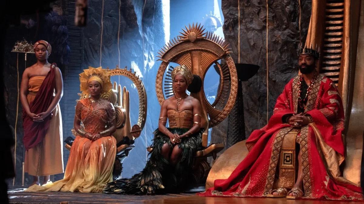 Four people in elaborate ceremonial costumes are arranged around ornate seats in a dramatic rocky chamber.
