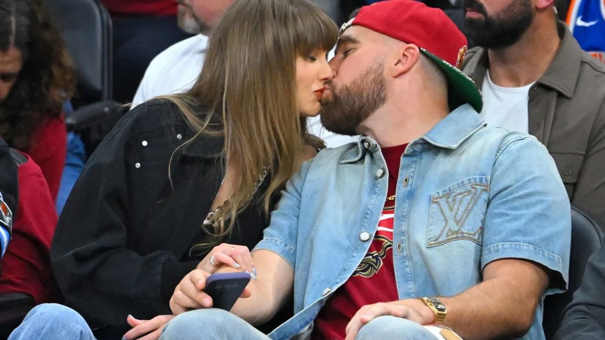 Two people kiss while seated among spectators; one wears a red cap and denim shirt.