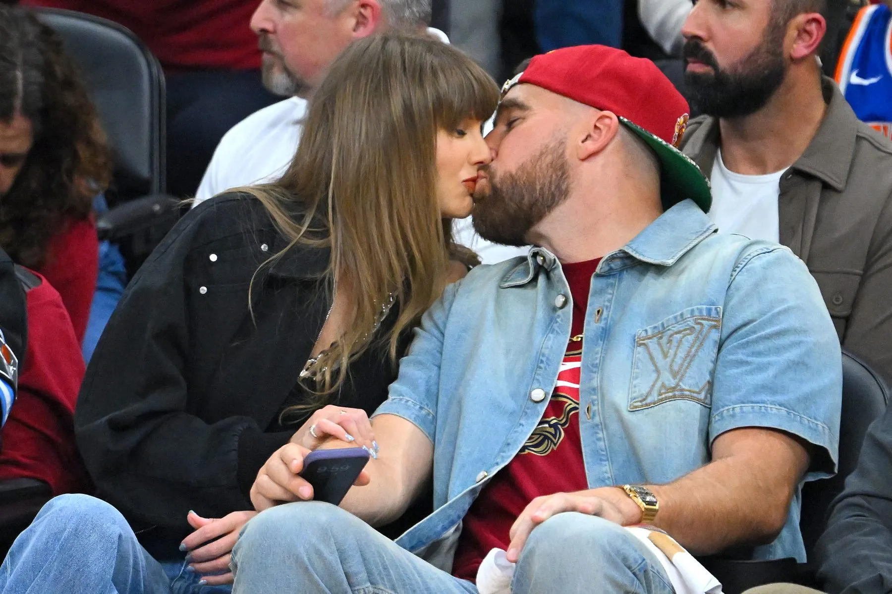 Two people kiss while seated among spectators; one wears a red cap and denim shirt.