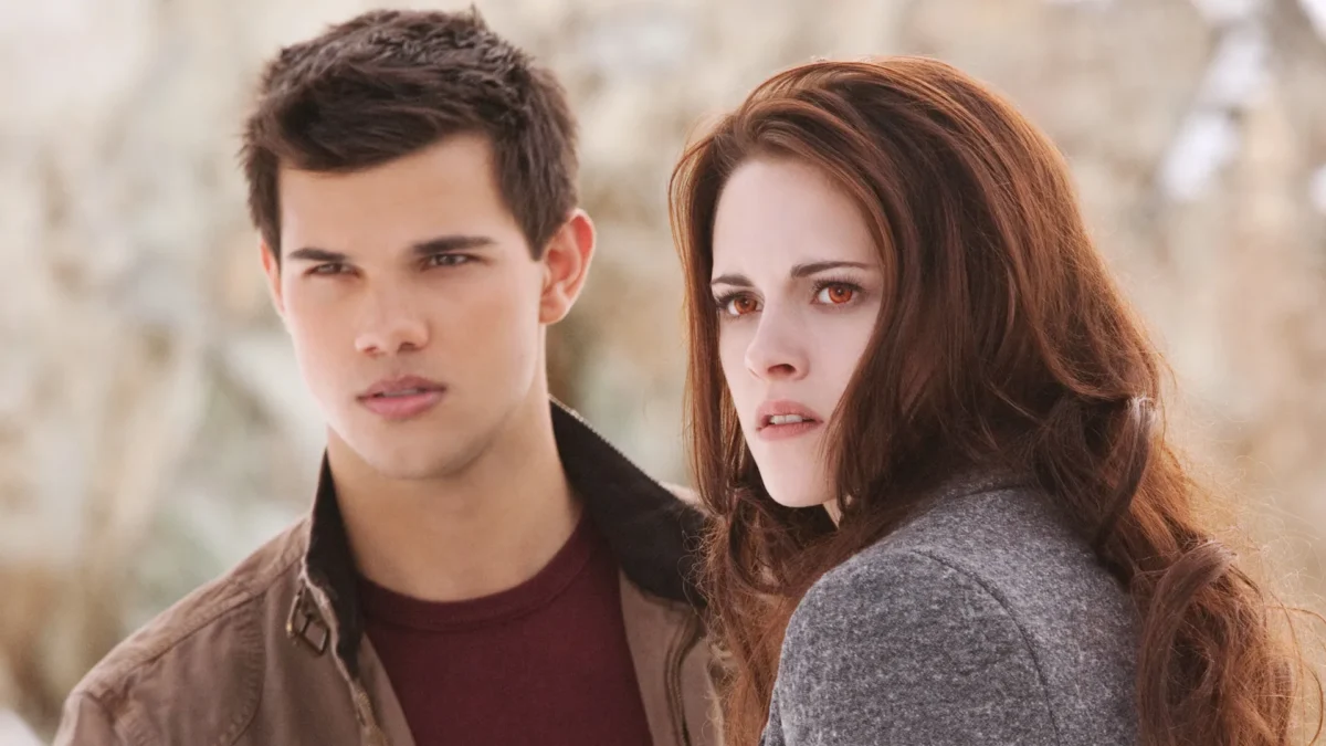 Two people, one with long reddish-brown hair, stare seriously off-camera against a blurred outdoor background.