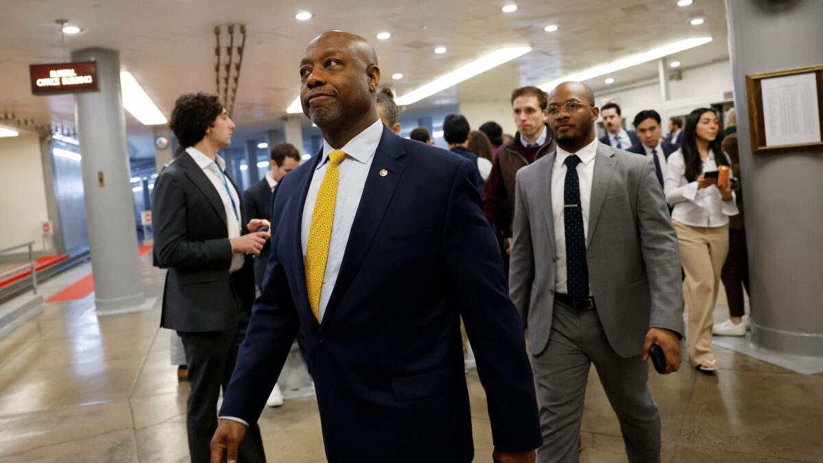 A man in a navy suit and yellow tie walks through a busy hallway with other suited people.
