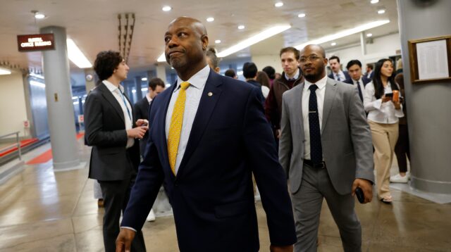 A man in a navy suit and yellow tie walks through a busy hallway with other suited people.