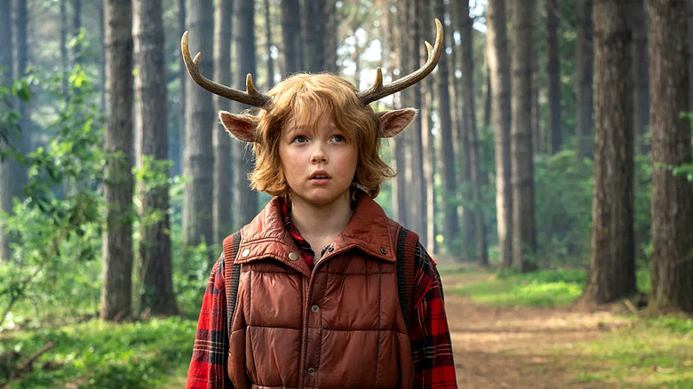 A child with deer antlers and ears stands on a sunlit forest path.