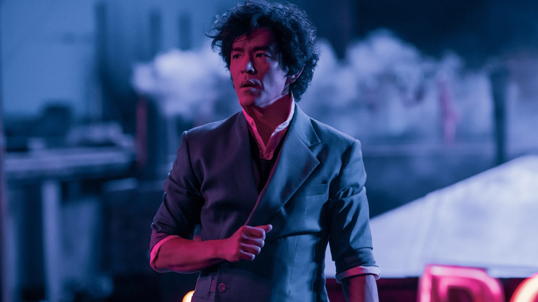 A man in a gray suit stands under blue and pink lighting, looking alert.