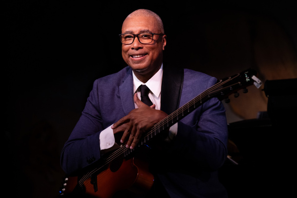 A smiling man in a blue suit and glasses holds a guitar against a dark background.
