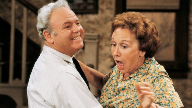 A smiling man in a white shirt holds a surprised woman in a floral dress indoors.